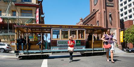 Cable car on San Francisco street with passengers boarding and alighting near historic brick buildings.