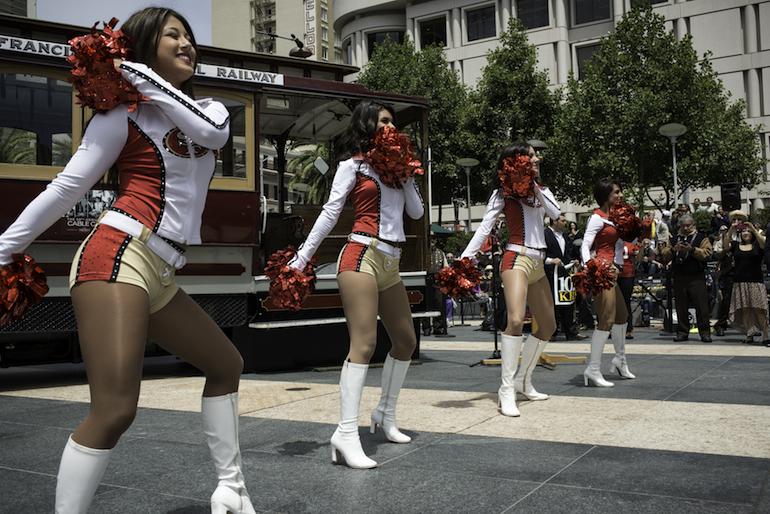 Cheerleaders in red and white uniforms with pom-poms dancing in front of a cable car at Union Square.