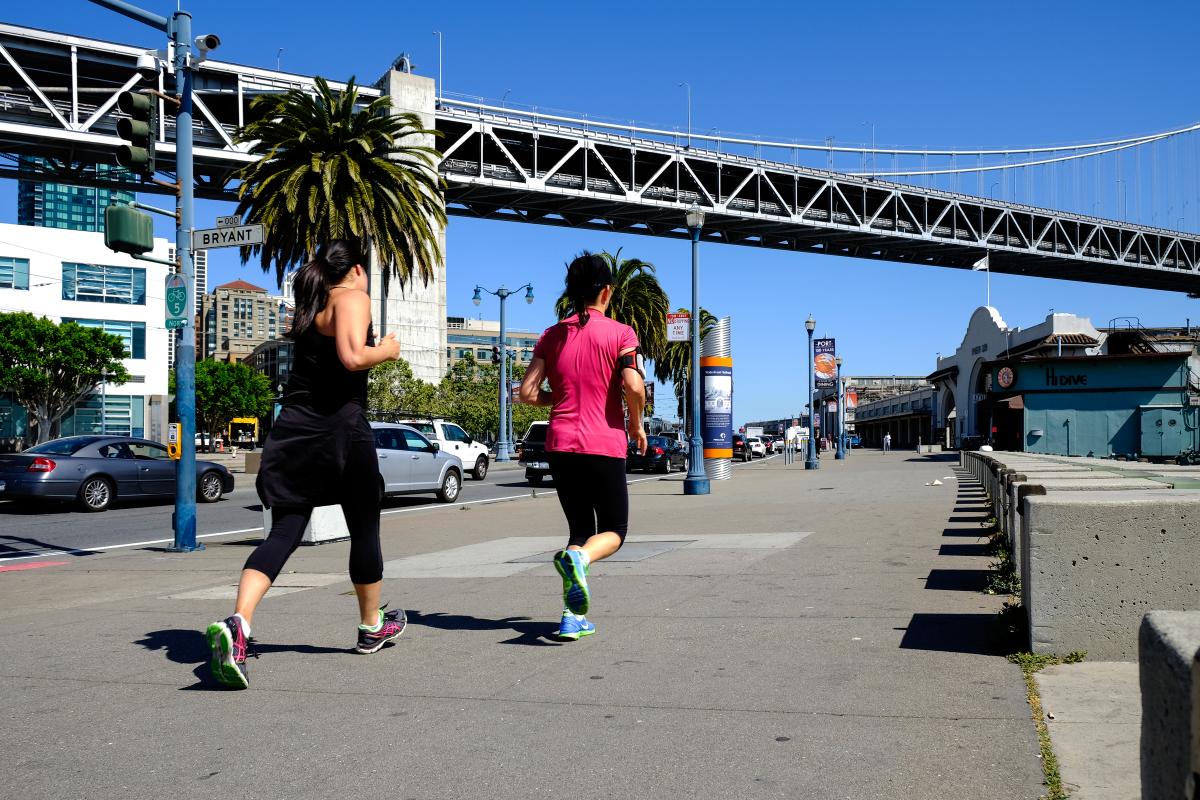 Two women jogging along the Embarcadero under the Bay Bridge on a sunny day.