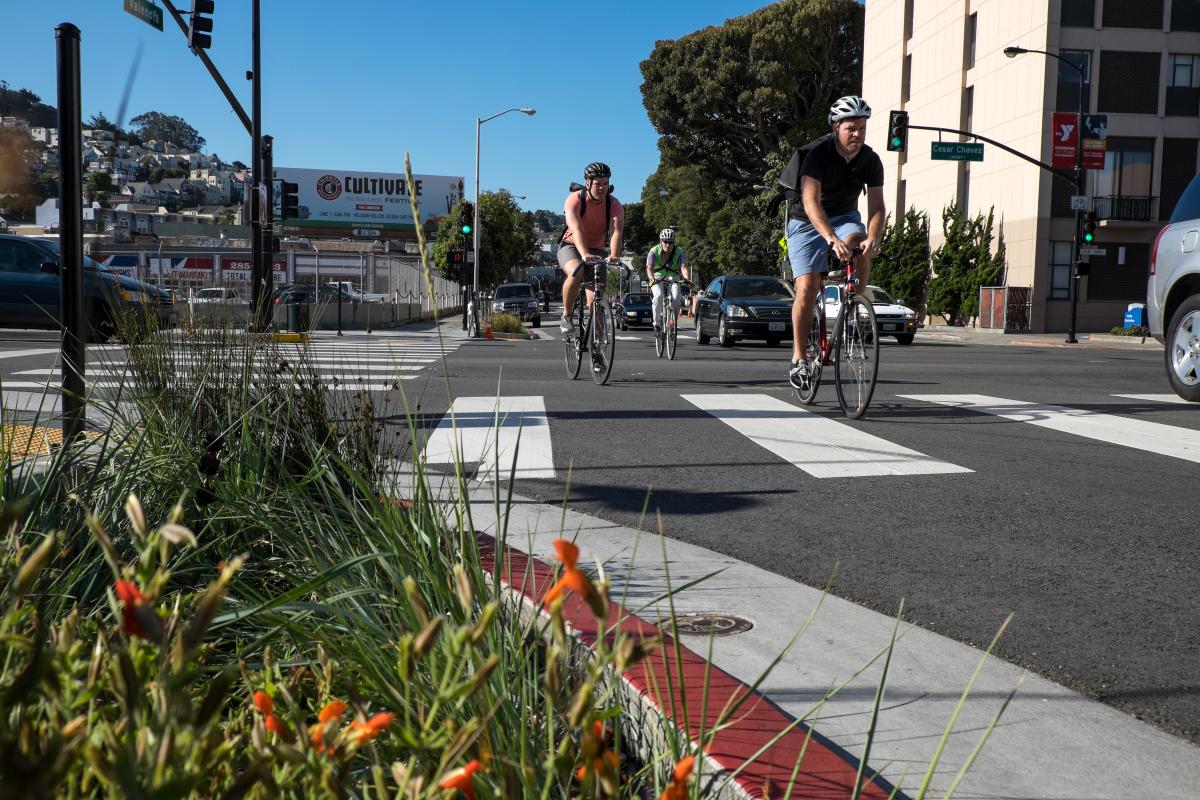 Cyclists and cars crossing Cesar Chavez Street at Valencia intersection with orange flowers in foreground.