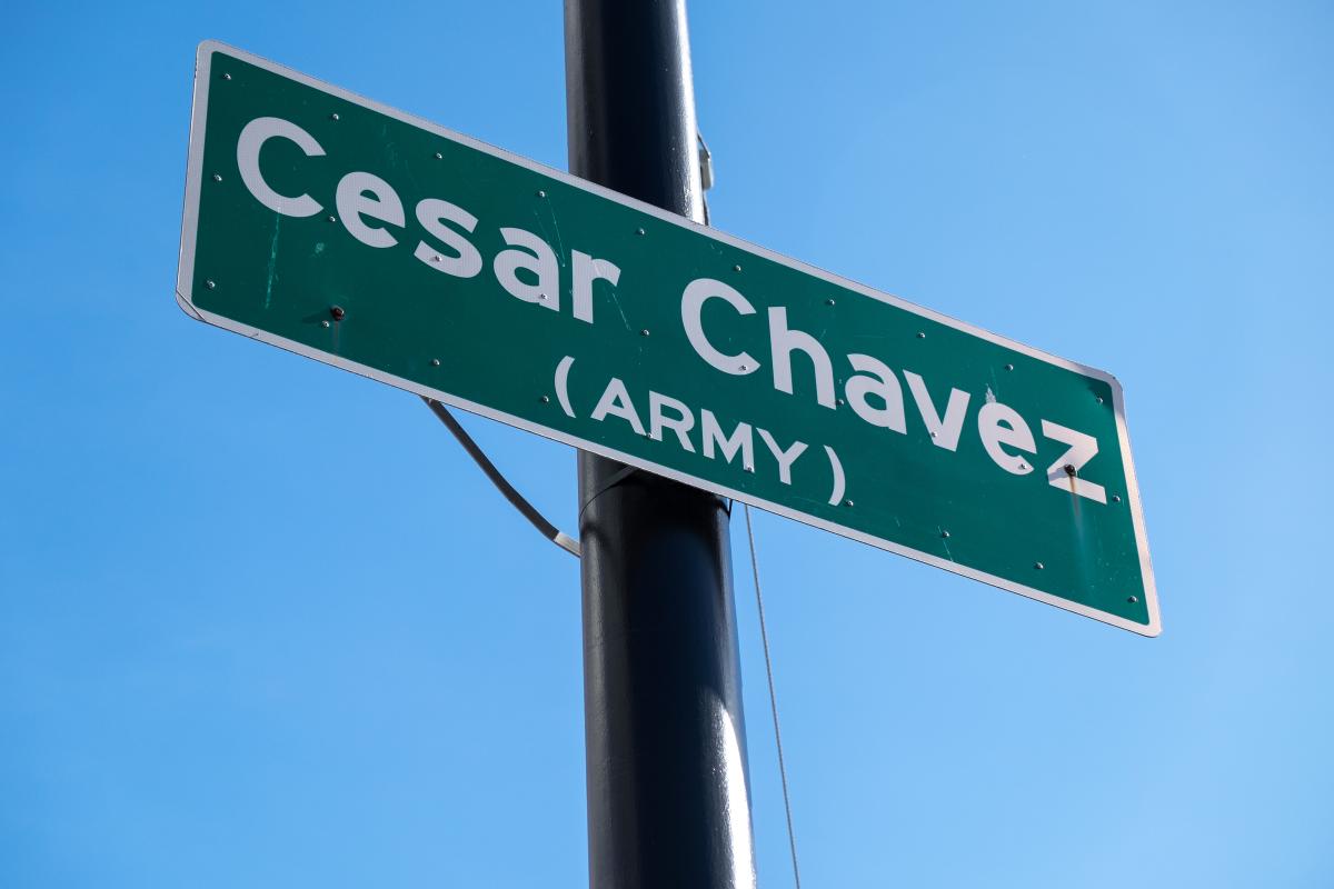 Green street sign reading "Cesar Chavez (ARMY)" mounted on a black pole against clear blue sky.