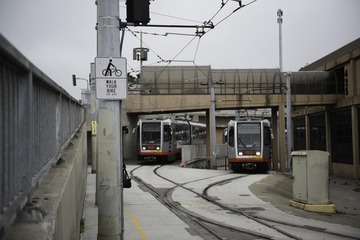 Two light rail vehicles on curved tracks at Balboa Park Station with overhead wires and a walk your bike sign visible.