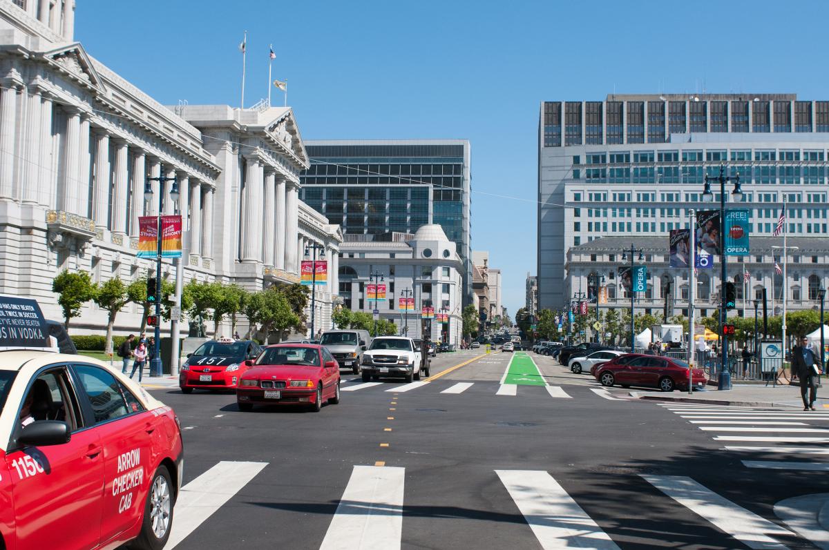 Vehicles and taxis on a wide street at San Francisco's Civic Center with classical and modern buildings, bike lane, and crosswalks visible.