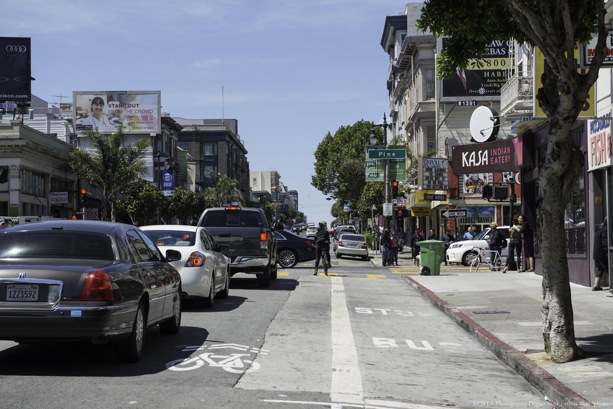 Busy urban street with parked and moving cars, pedestrians, storefronts, and large trees lining the sidewalk.