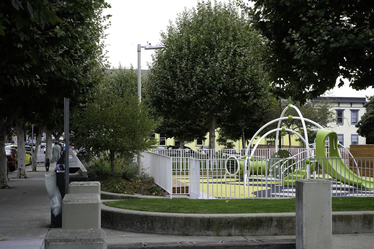 Children's playground with green and white play equipment behind white fencing on a tree-lined street.
