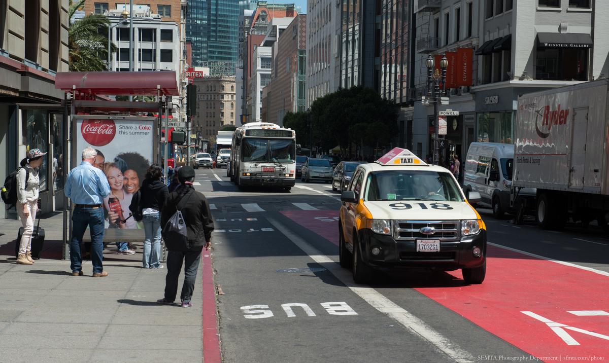 38 Geary bus approaches passengers on Geary at Powell, sharing red transit lane with yellow taxi and delivery trucks.