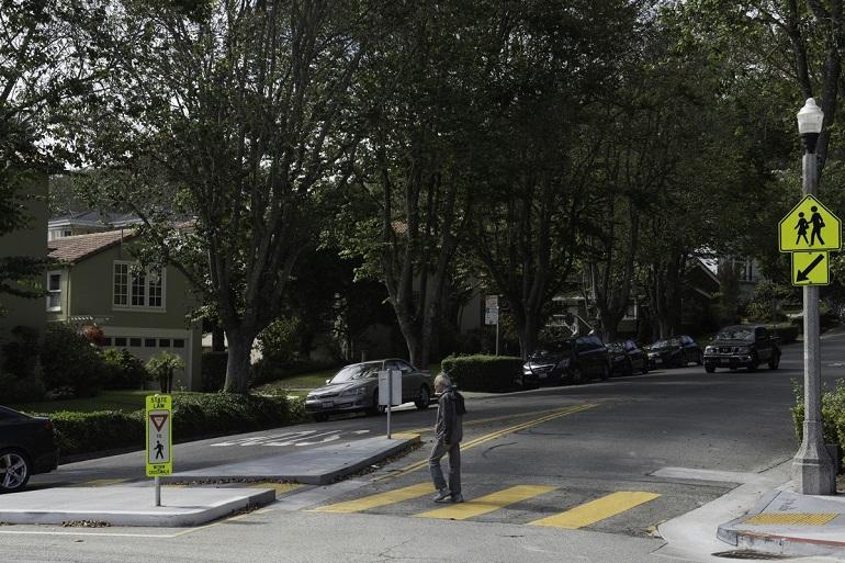 Pedestrian crossing a street at a marked crosswalk with a concrete island, near residential homes and trees.