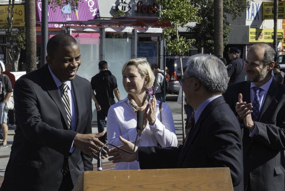 Four officials in business attire at a downtown street press conference, with palm trees and storefronts visible behind them.