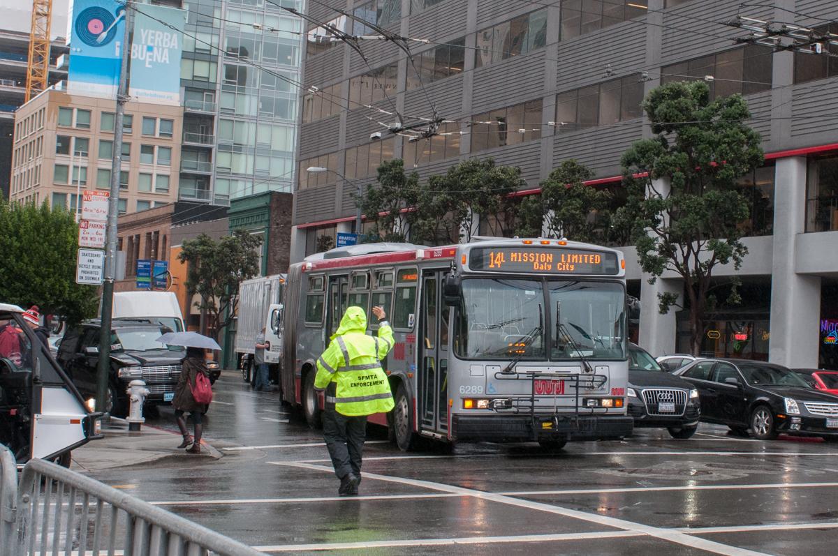 Parking Control Officer in yellow rain jacket directs 14L Mission Limited bus through wet Mission Street traffic.