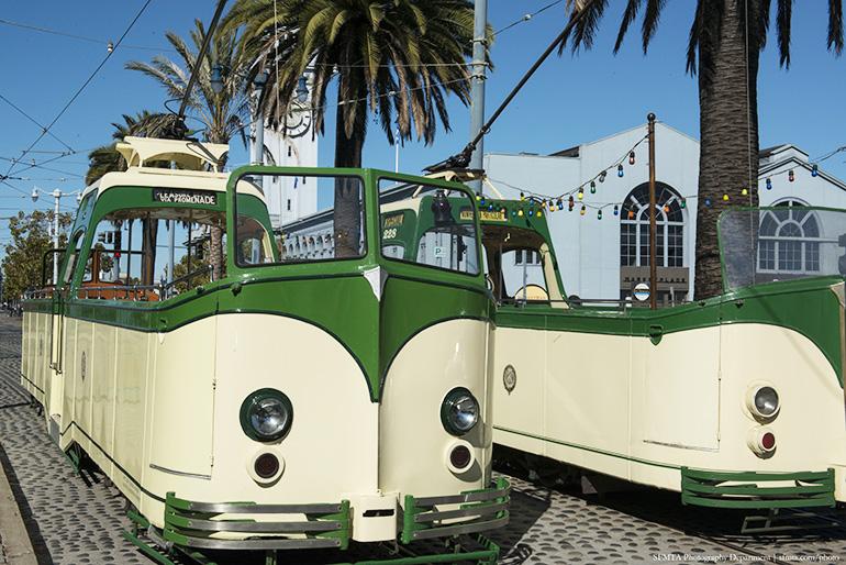 Two green and cream vintage streetcars parked in front of the Ferry Building with palm trees and blue sky.