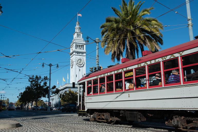 Red and white historic streetcar passing Ferry Building clock tower under clear blue sky with palm tree.