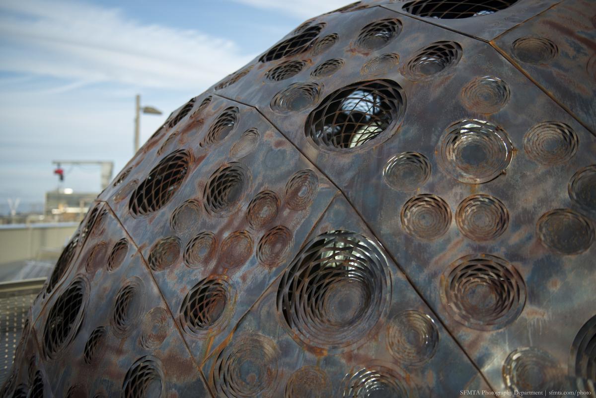 Close-up of rusted steel sculpture with circular perforations and openings, waterfront visible in background.