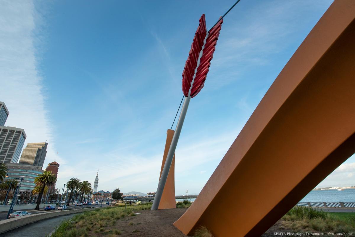 Large red arrow sculpture on waterfront with city skyline and bay in background