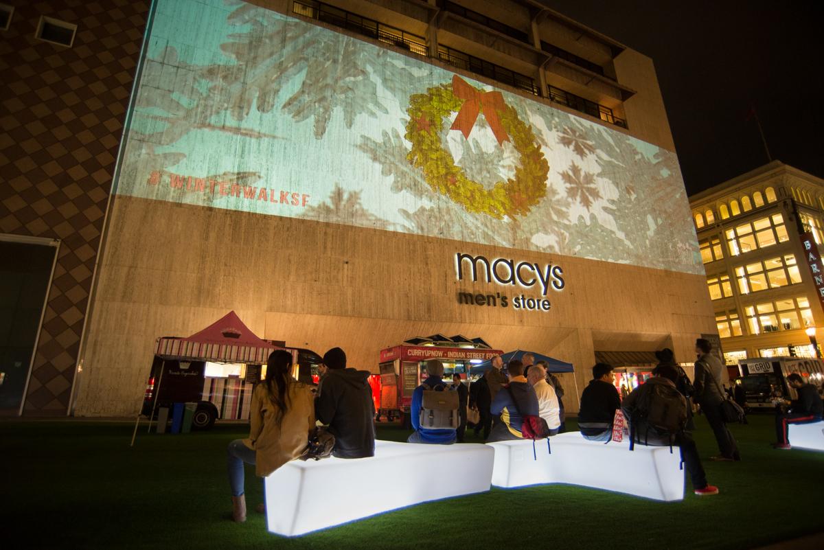 People gather on a plaza with illuminated benches watching a holiday light projection on a Macy's building facade.