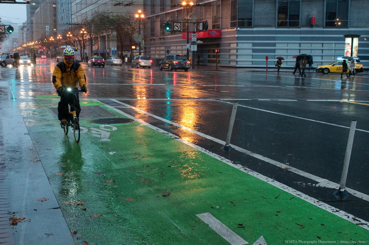 Cyclist in yellow jacket riding on green-painted bike lane during rainy evening on Market Street.