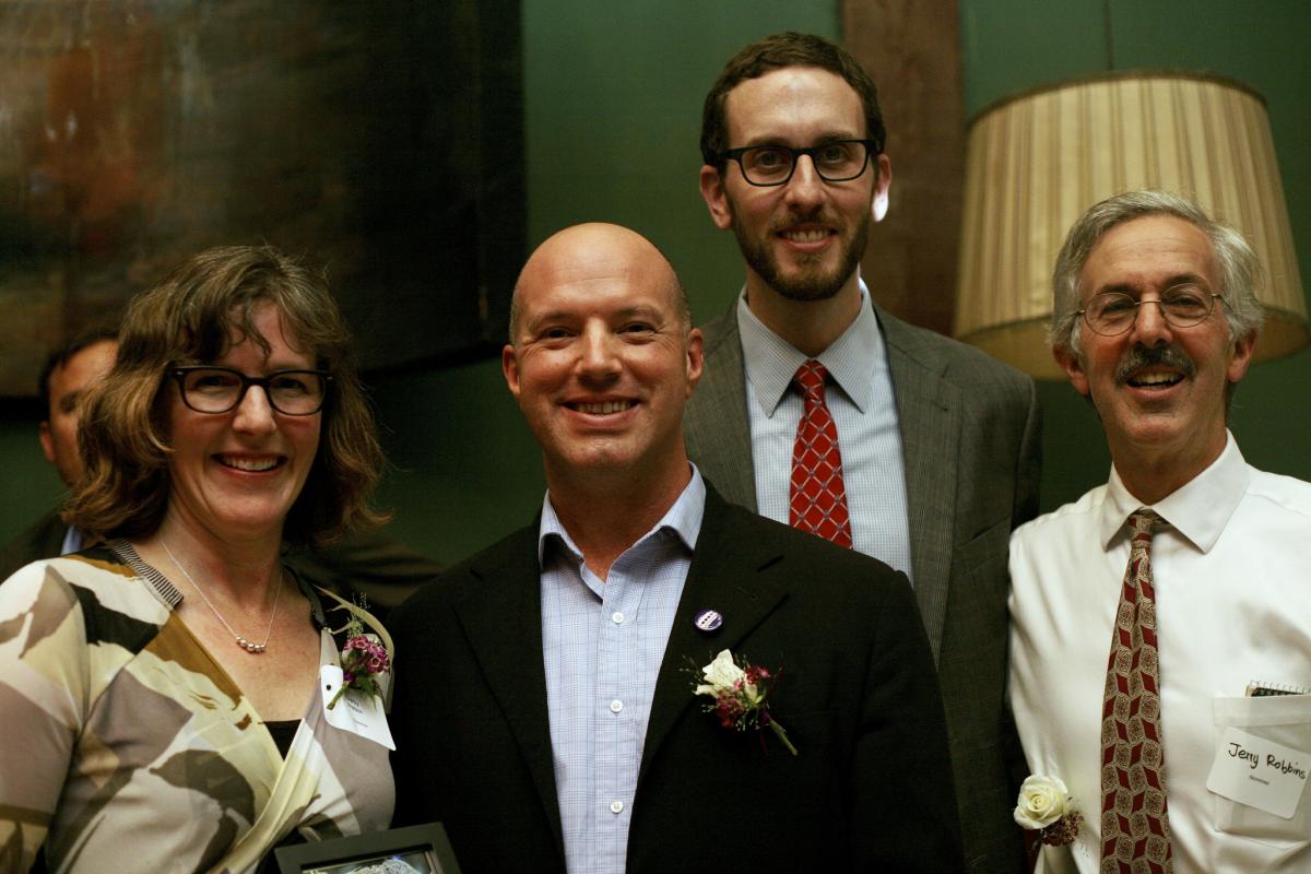 Four people in business attire smiling at an indoor event, with one wearing a boutonniere.