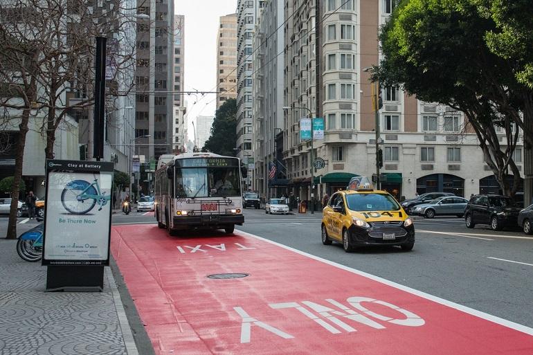 Muni bus in red transit-only lane on Clay Street with taxi and bike-share station visible.