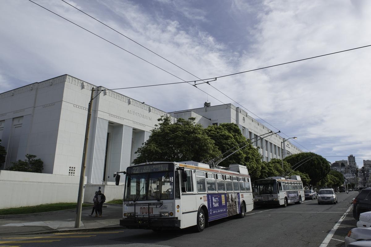 White transit buses lined up on a street in front of a large white building with overhead power lines.