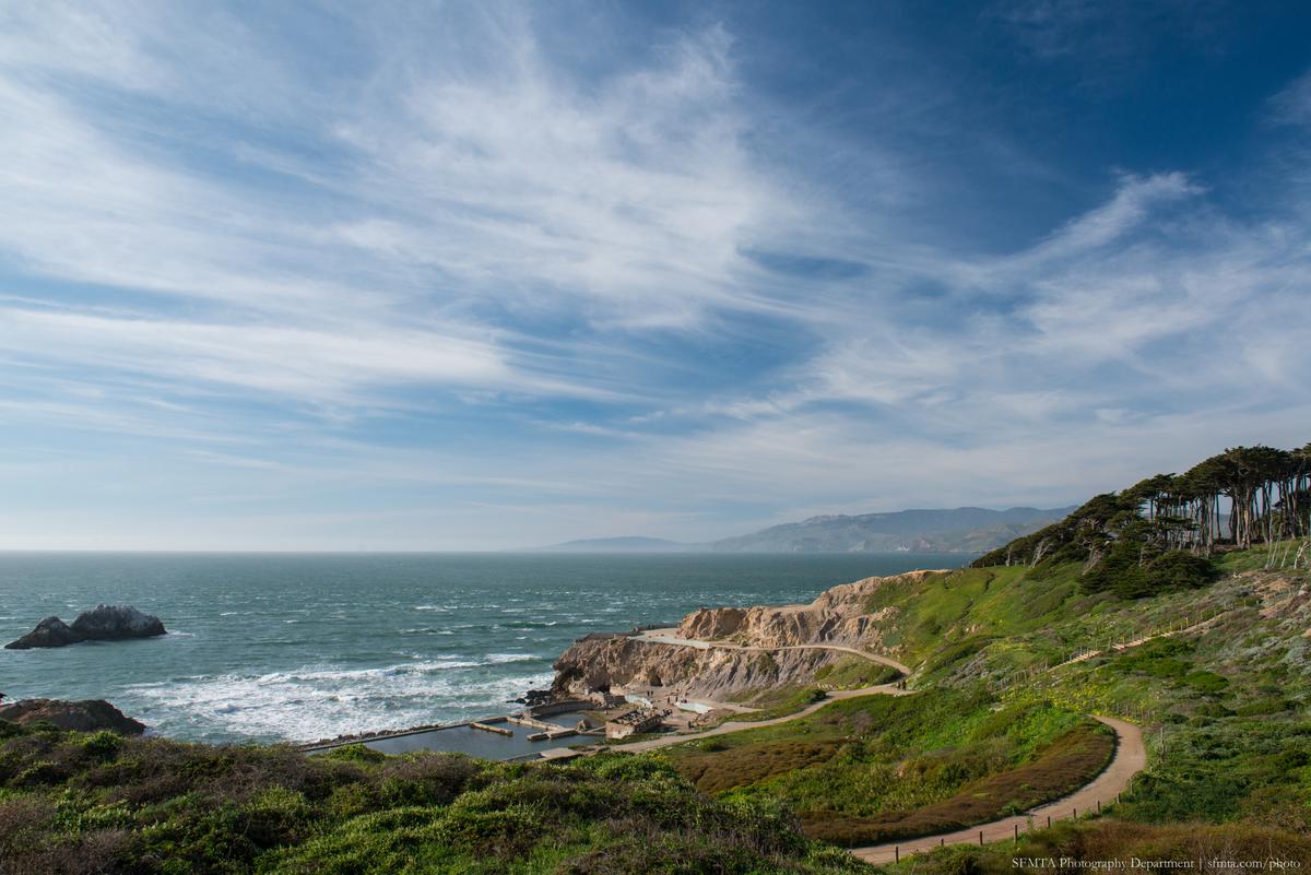 Coastal cliffside path overlooking rocky shores and ocean near San Francisco with cypress trees and mountains in distance.