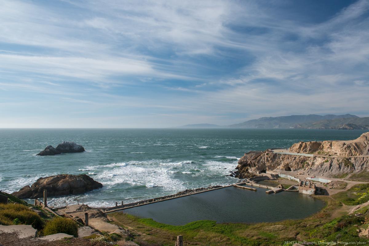 Sutro Baths ruins with ocean, rocky cliffs, and distant hills under blue sky with clouds.