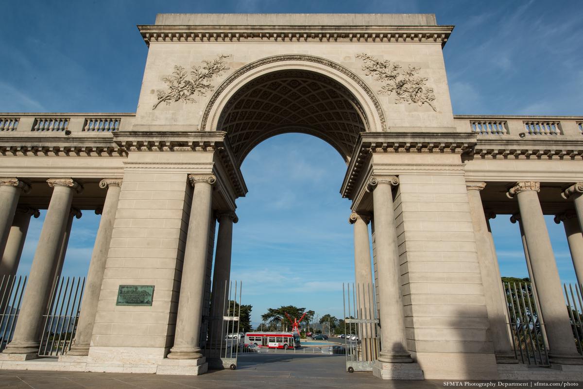 Neoclassical stone archway with columns and a red Muni bus visible through the opening against a blue sky.