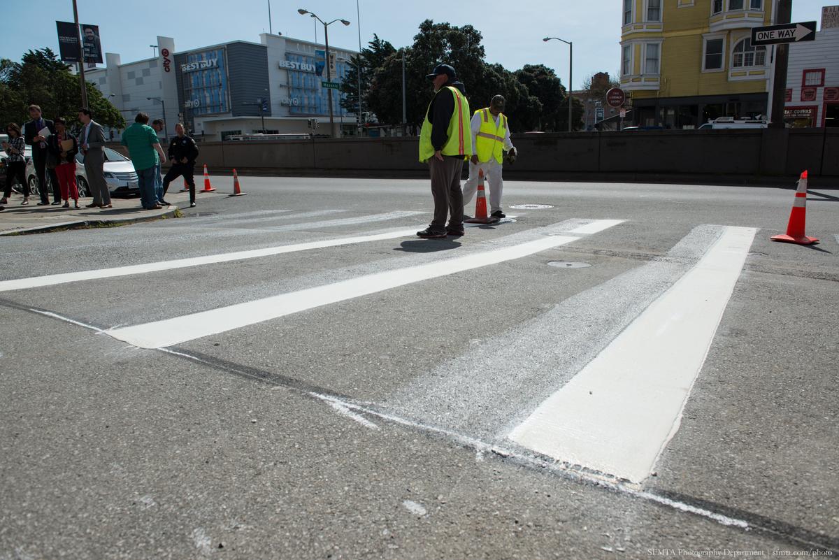 Newly painted zebra-striped crosswalk at intersection with workers in safety vests and traffic cones.