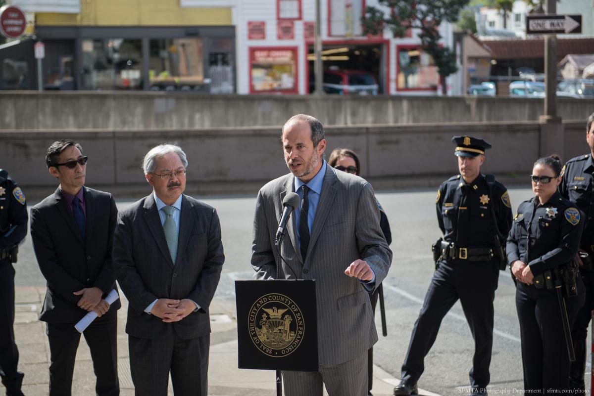 Man in pinstriped suit speaks at podium with city seal while officials and police officers stand nearby on urban street.