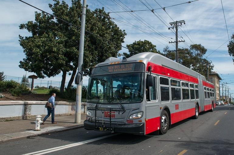 Red and silver articulated Muni bus stopped at curb with passenger waiting nearby.