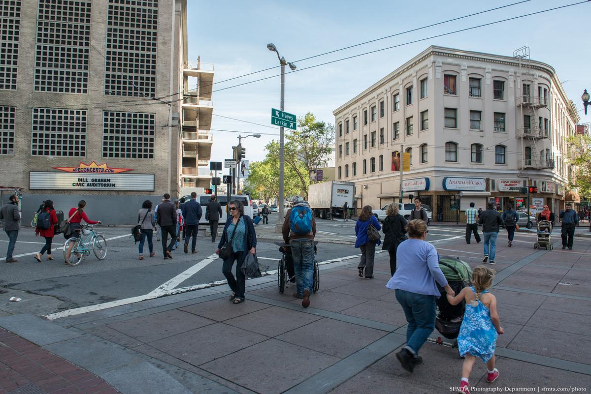 Pedestrians with strollers, a wheelchair and a bicycle traverse the intersection of Market, Hayes and Larkin streets.