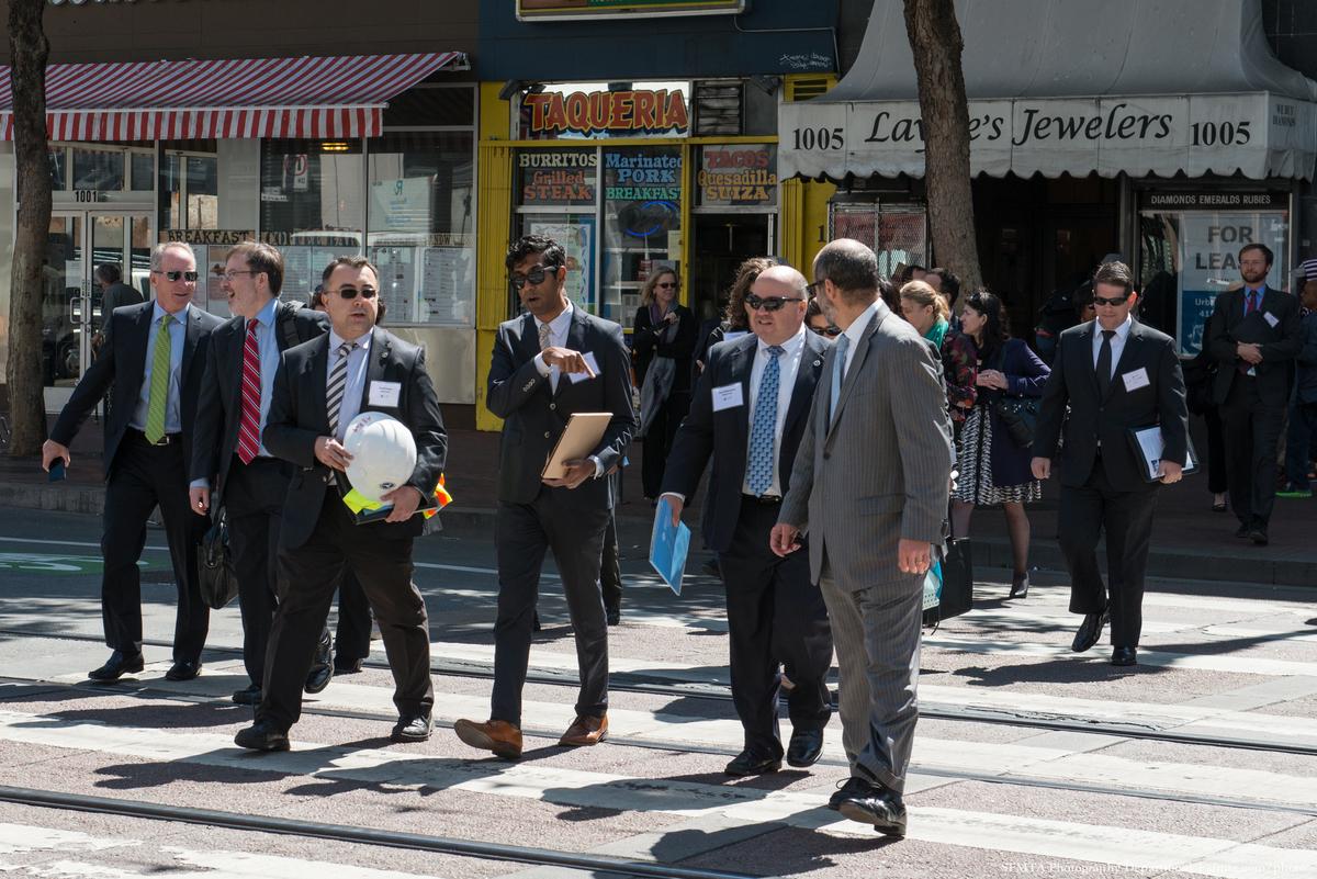 Group of business professionals in suits crossing a city street at a marked crosswalk with storefronts visible behind them.