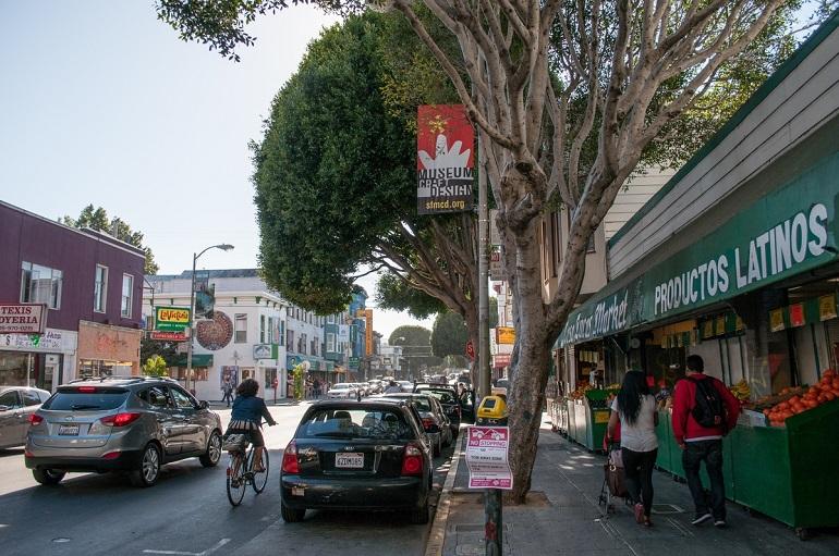 Neighborhood street with storefronts, pedestrians, cyclist, and parked cars under large trees.