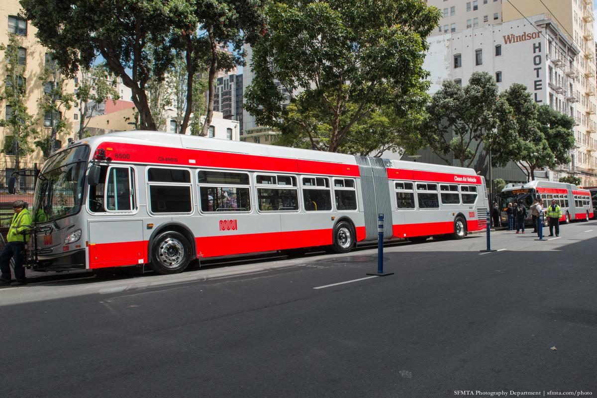 Red and white articulated bus parked on city street with buildings and trees in background.