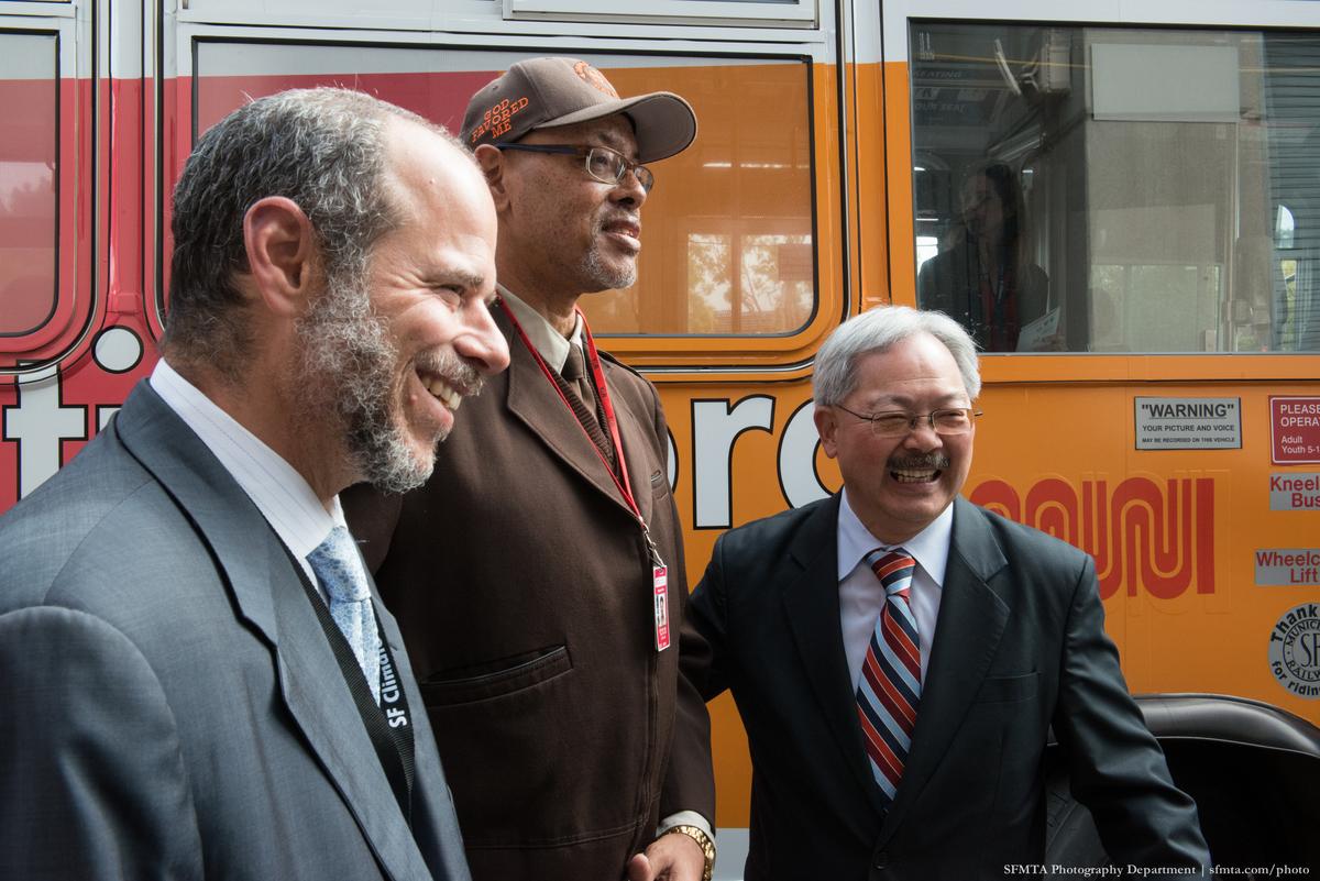 Three men in business attire stand in front of an orange and red transit bus, smiling at a public event.
