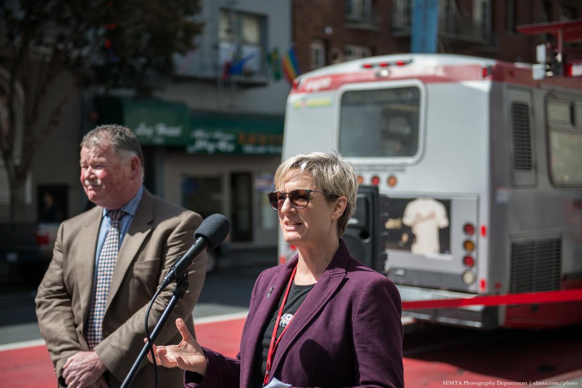 Two people speaking at a podium in front of a transit bus on a city street.