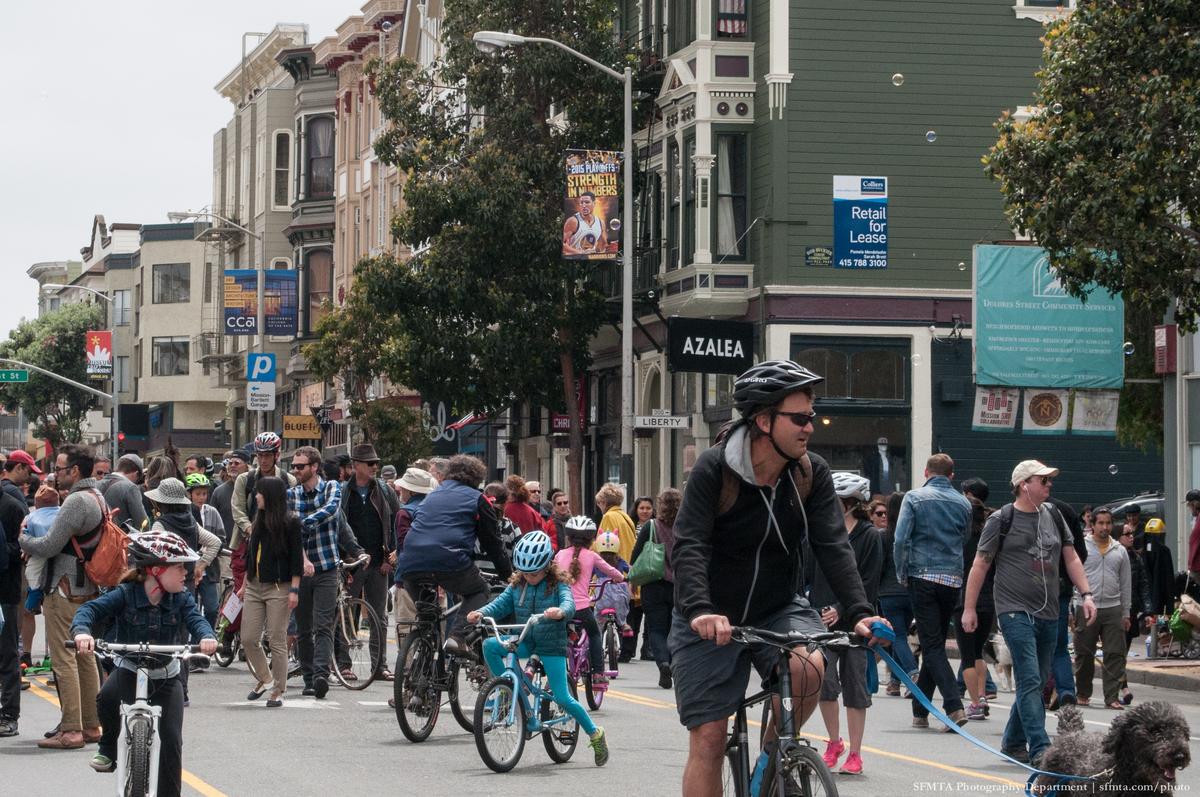 Cyclists and pedestrians fill a closed urban street lined with historic buildings and trees during Sunday Streets event.