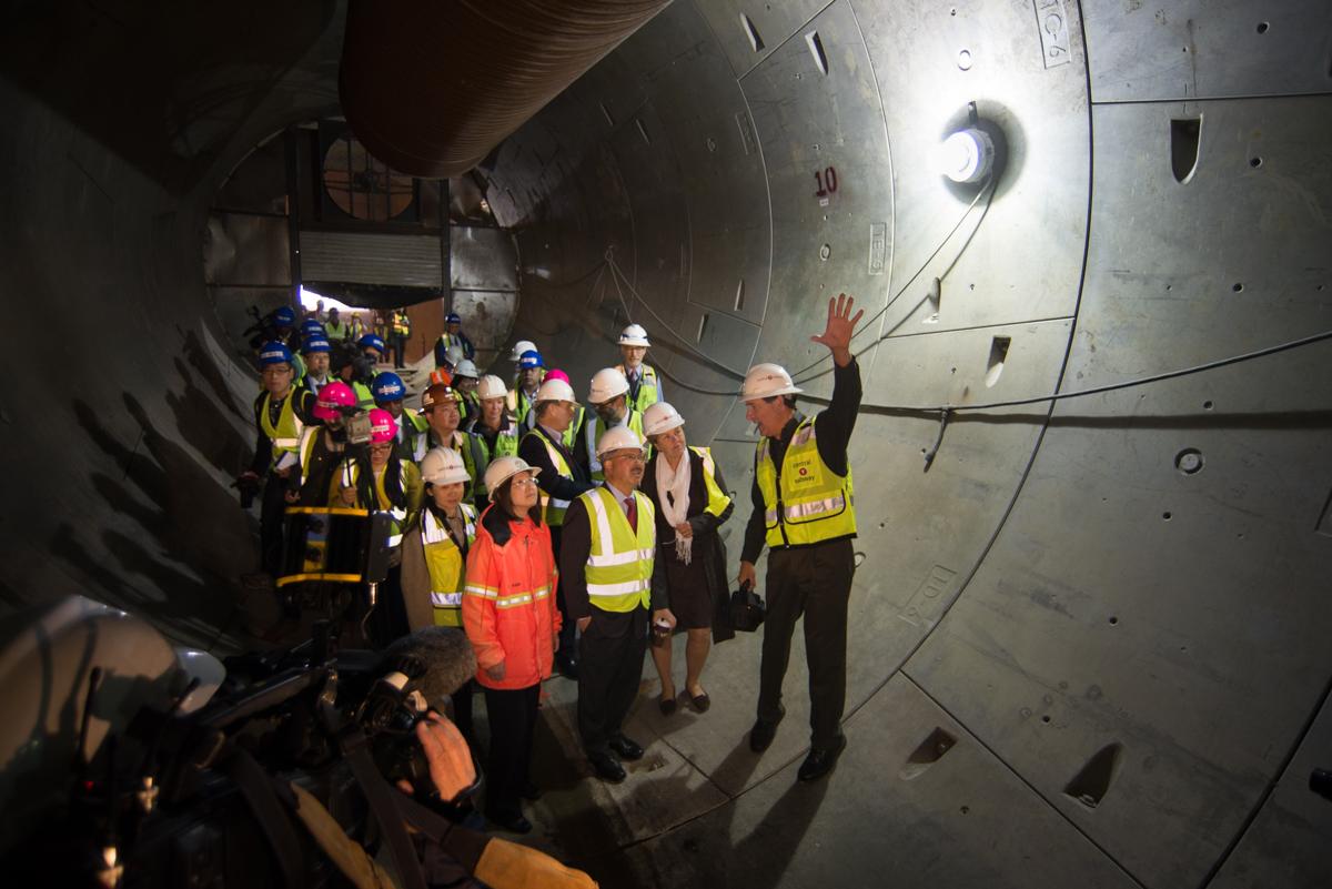 Group of officials and media wearing hard hats and safety vests inside a concrete subway tunnel under construction.