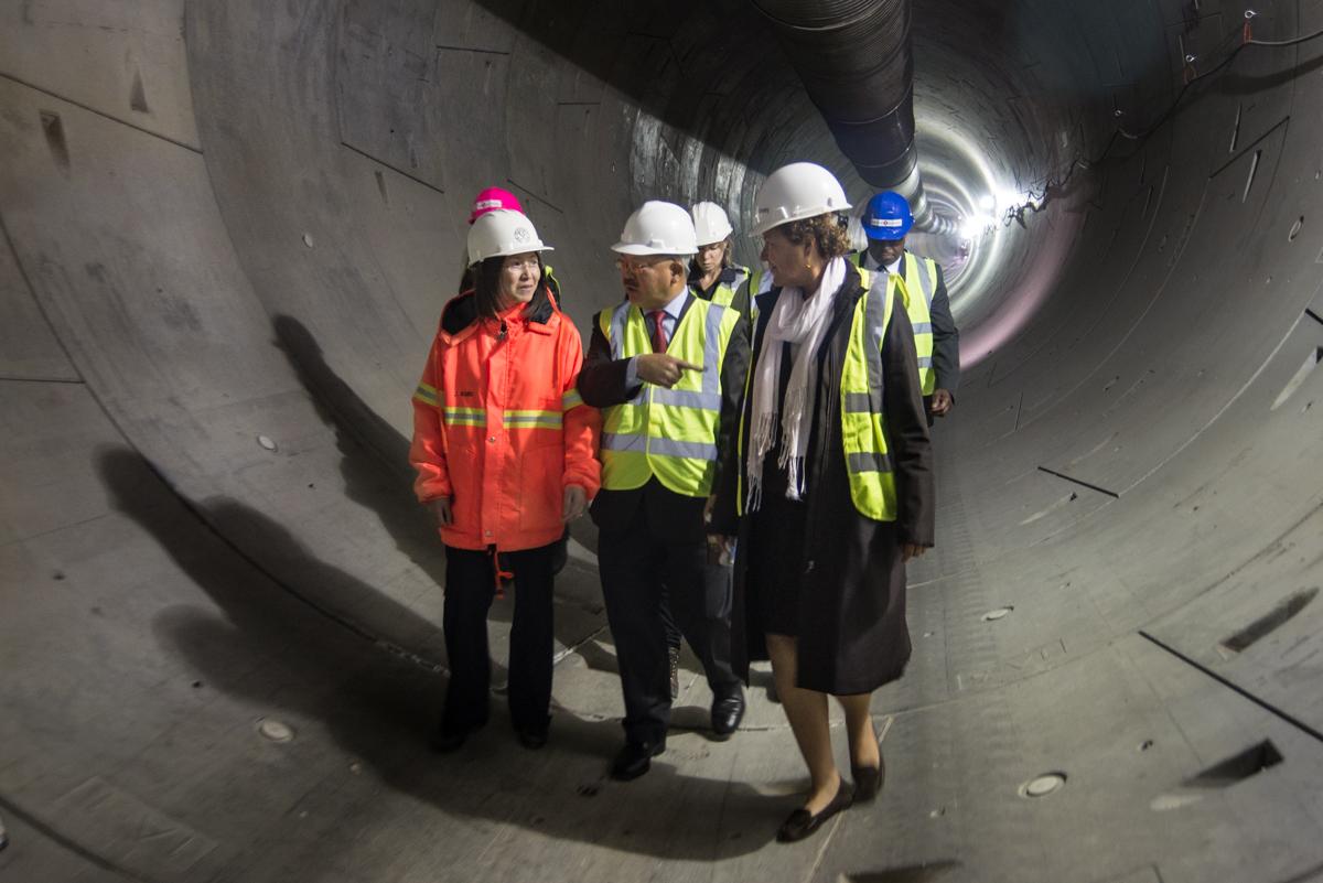Four construction workers in hard hats and safety vests tour a large concrete tunnel under construction.