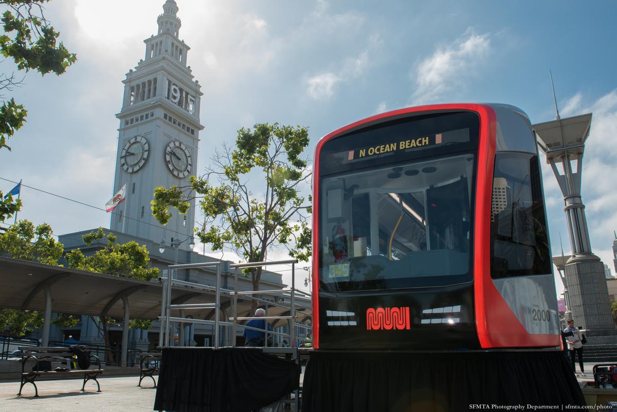 Red Muni light rail vehicle mock-up displayed on Embarcadero plaza with Ferry Building clock tower in background.