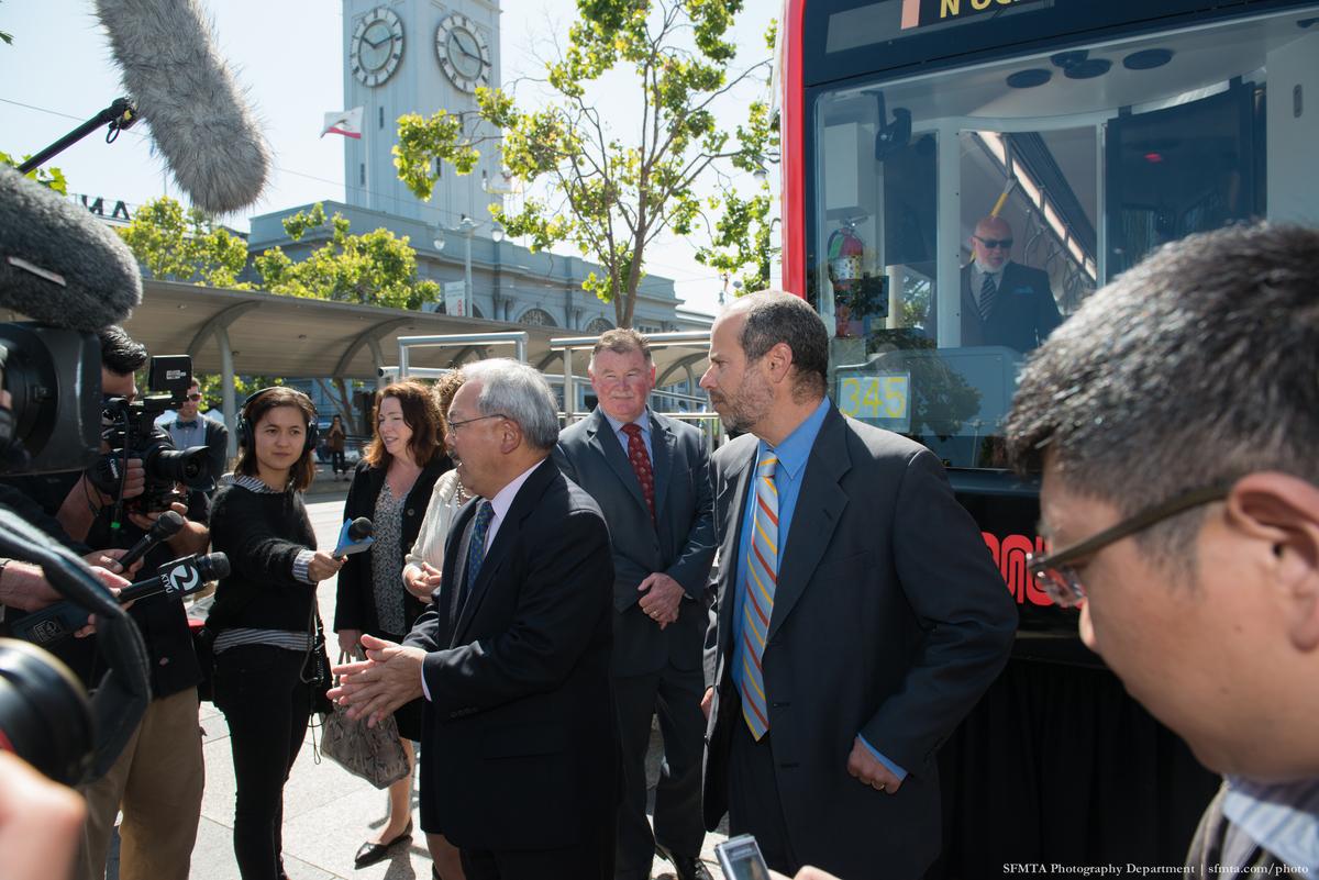 Mayor Lee and officials speak to reporters at LRV mock-up event with media present and clock tower visible.