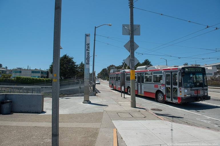Red and white Muni bus stopped at Balboa Park Station with overhead wires and station signage visible.