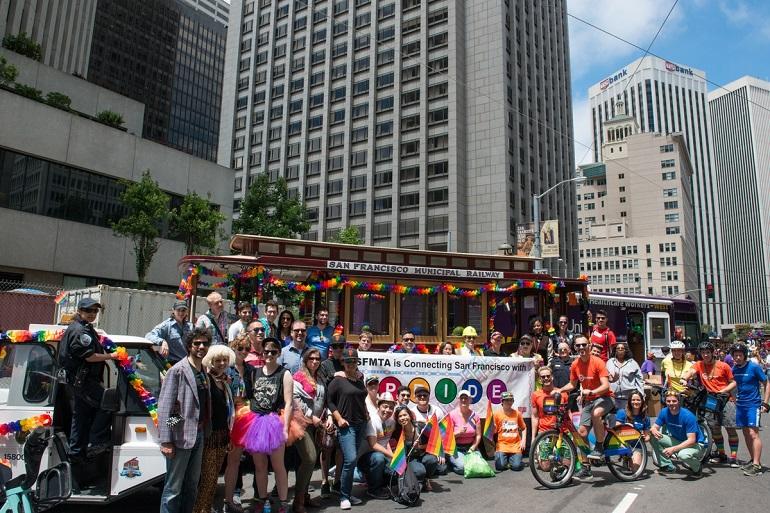 SFMTA cable car decorated with rainbow colors surrounded by Pride Parade participants and spectators in downtown San Francisco.