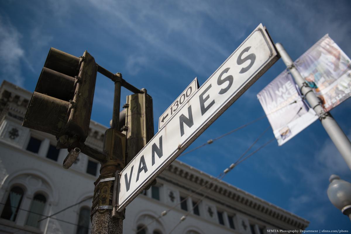 Van Ness Avenue street sign against blue sky with historic buildings and traffic light visible.