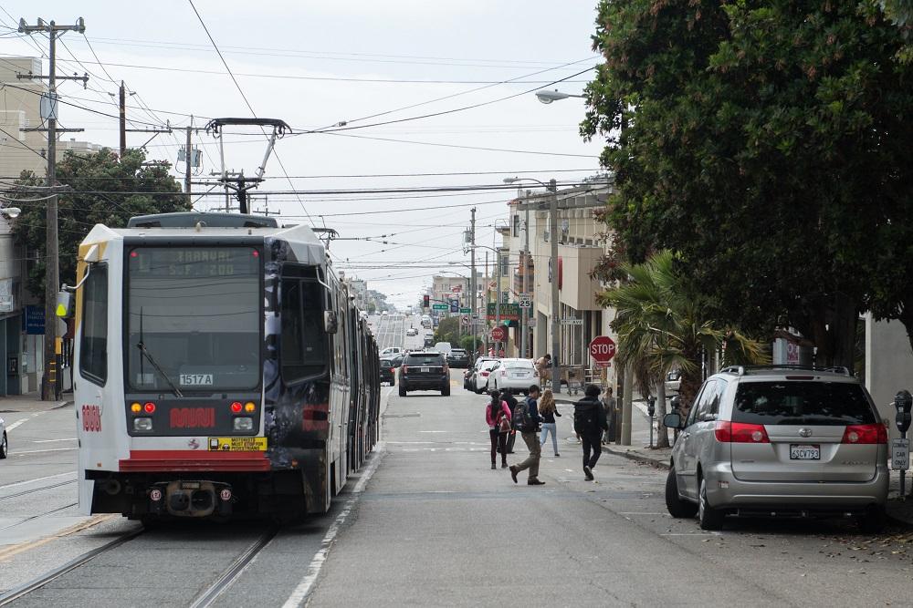 L Taraval light rail train stopped on street with pedestrians and cars nearby
