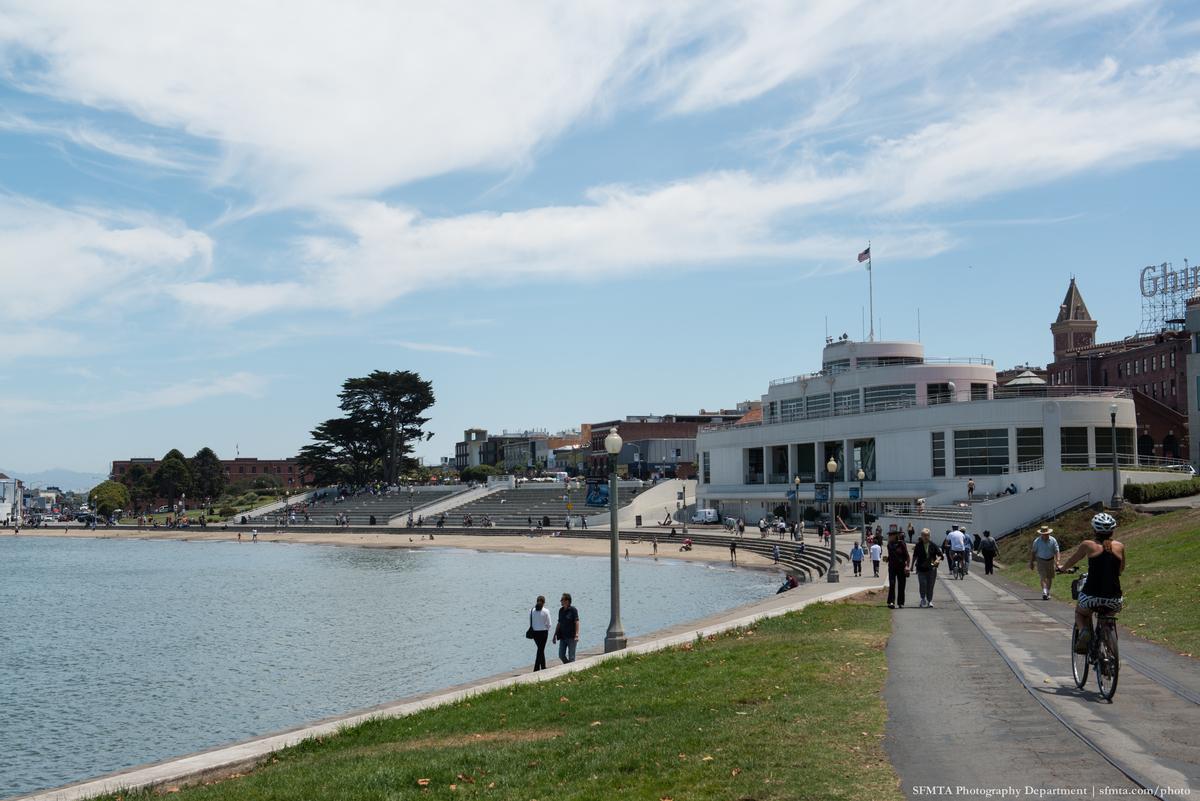 Waterfront promenade with white building, beach, and grassy area on sunny day