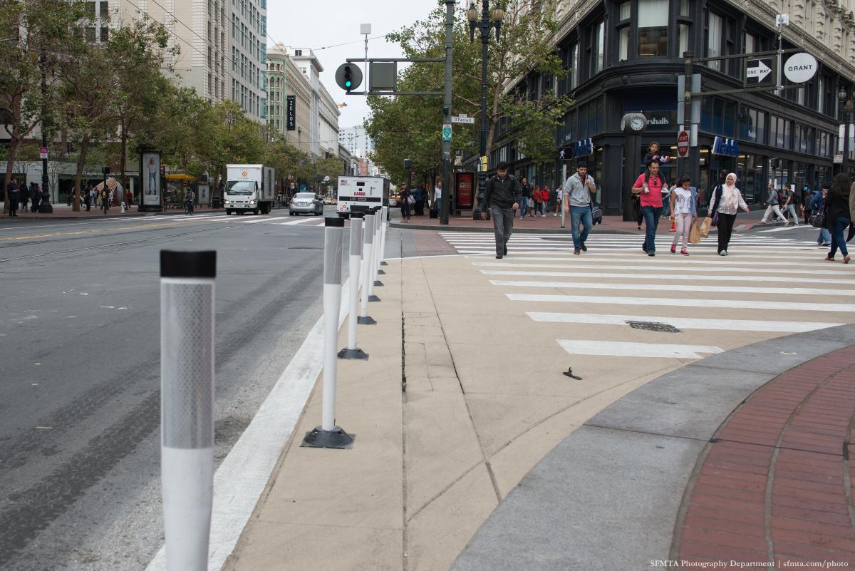 Pedestrians crossing at O'Farrell and Market with white protective posts and beige buffer zone along street.