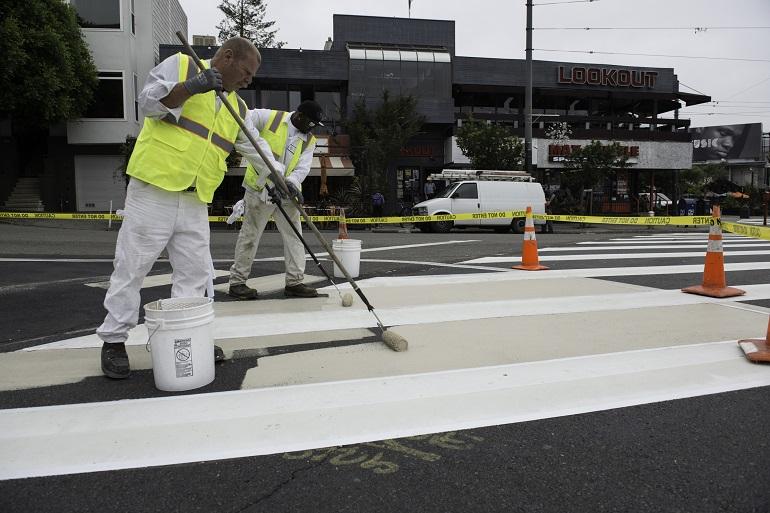 SFMTA worker in safety vest applying white paint to street crosswalk with roller tool.