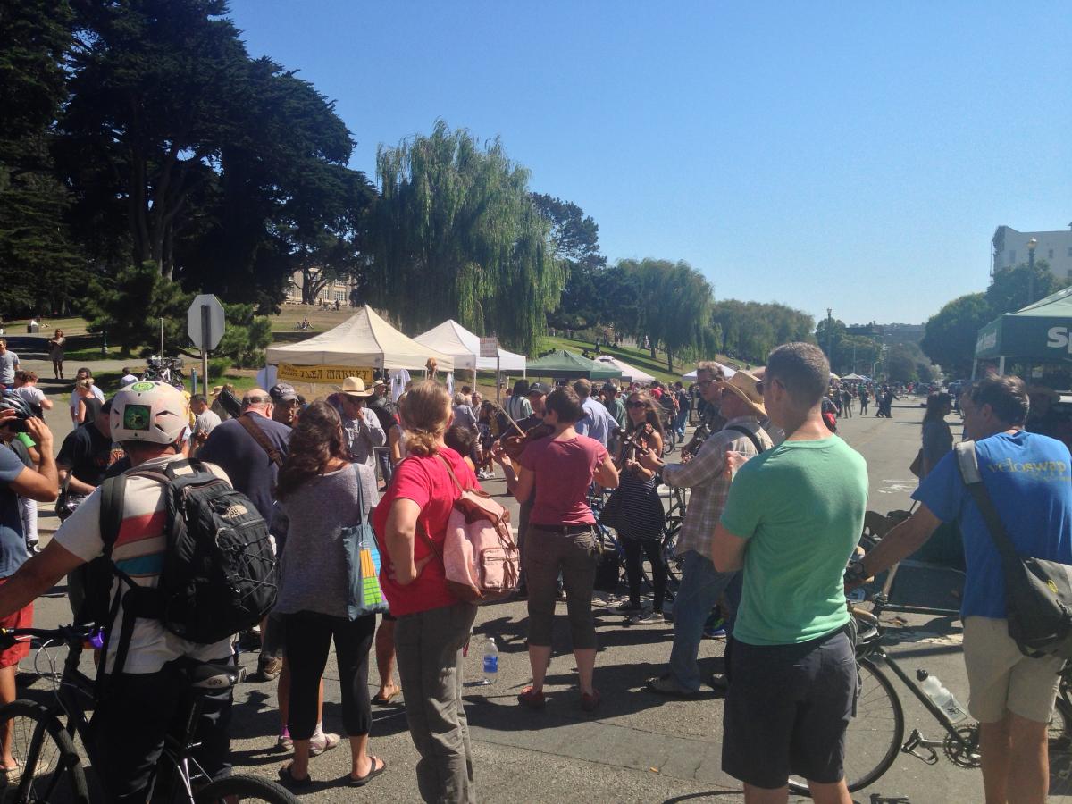 Crowd of people at outdoor market with tents, trees, and bicycles on sunny day