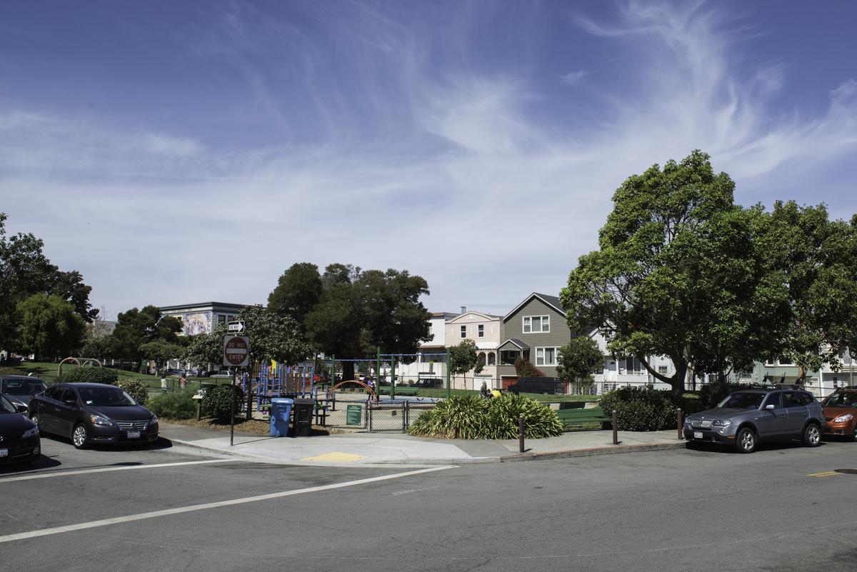 Precita Park with playground, trees, residential buildings, parked cars, and clear blue sky in Bernal Heights.