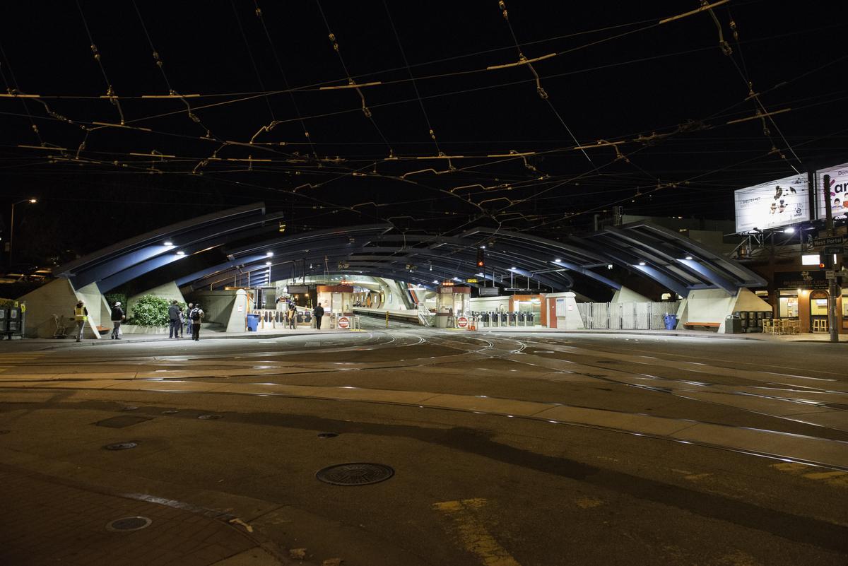 Night view of West Portal Station entrance with construction lighting and overhead power lines.