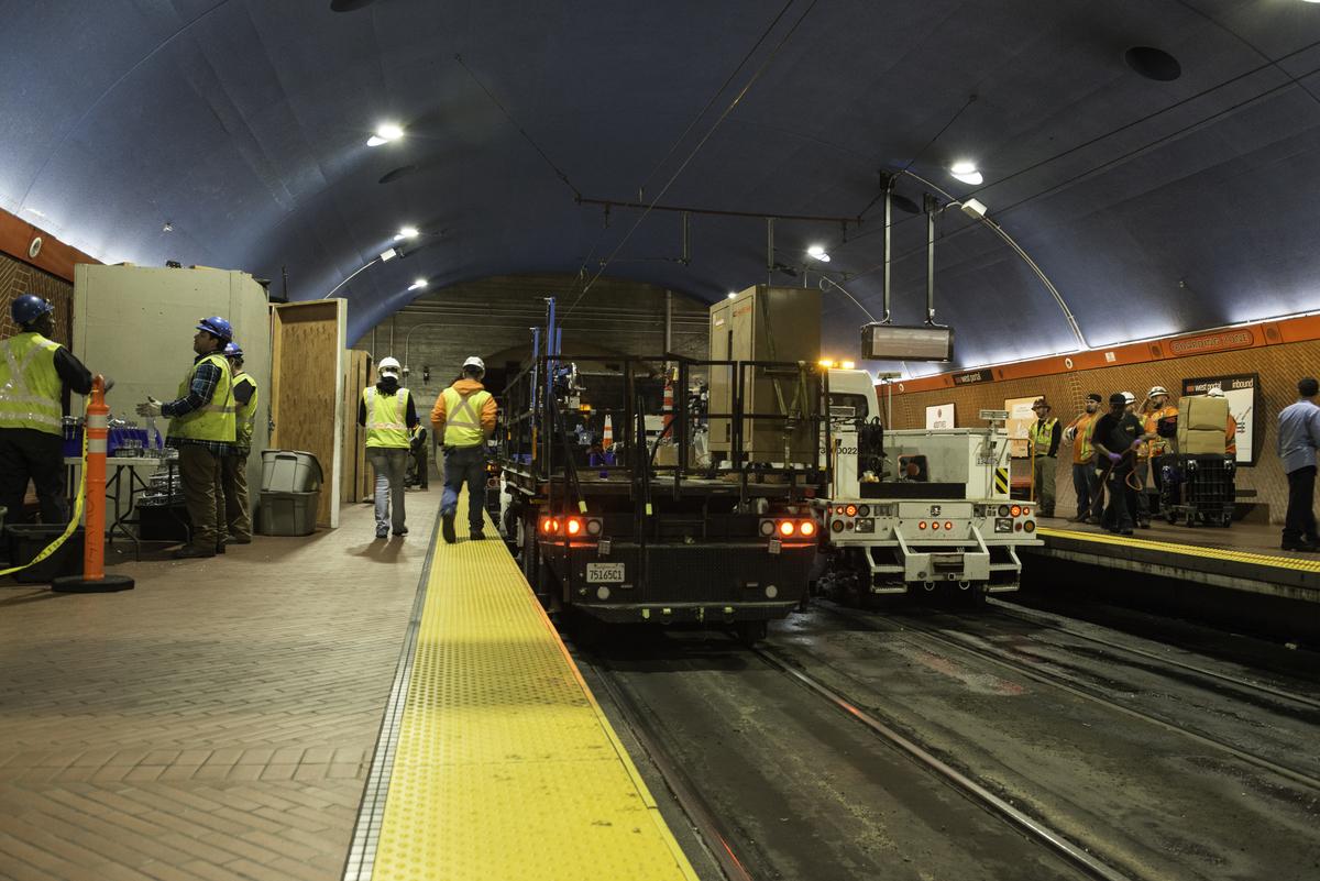 Workers in safety vests stand near a specialized rail truck inside West Portal Station tunnel during communications system installation.
